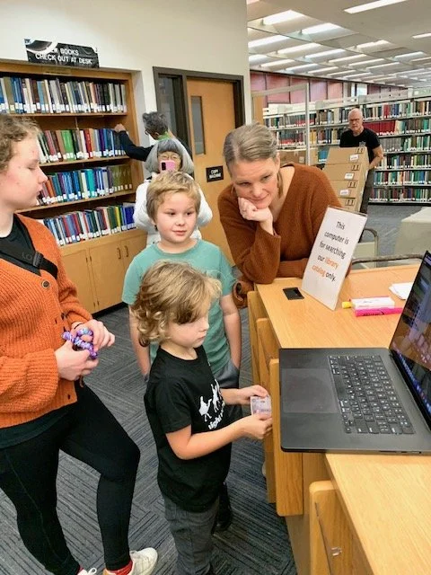 A woman and children in a library looking at a computer desk with a laptop. The woman appears to be explaining something, while the children listen attentively. Bookshelves are visible in the background.