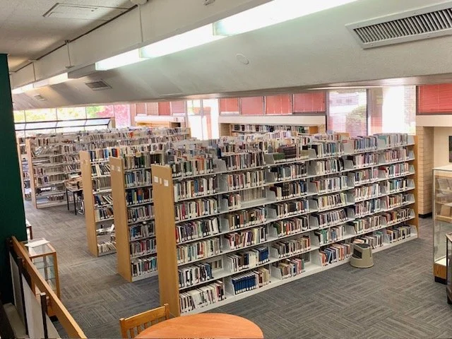 Interior of a public library with multiple shelves of books and a large window with natural light.