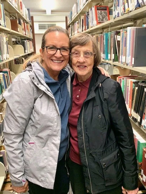 Two women smiling and hugging in a library aisle with bookshelves filled with books in the background.