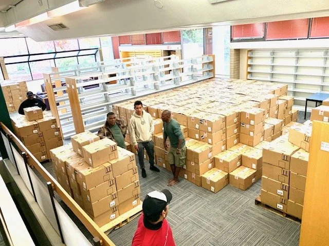 People inside a store surrounded by stacked cardboard boxes, some on display shelves, some on the floor, in a retail or warehouse setting.