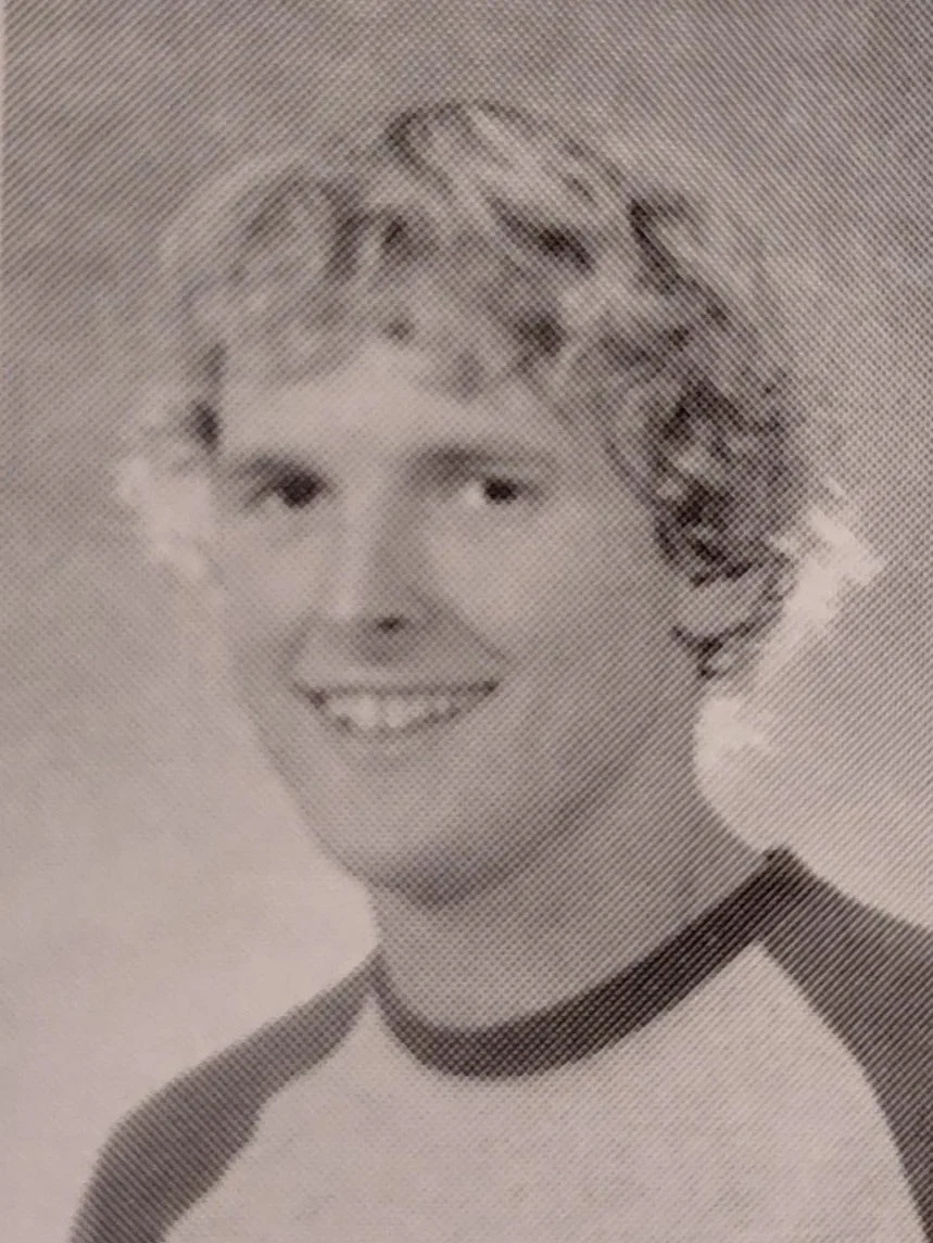 A young boy with curly hair, smiling and wearing a baseball-style shirt.