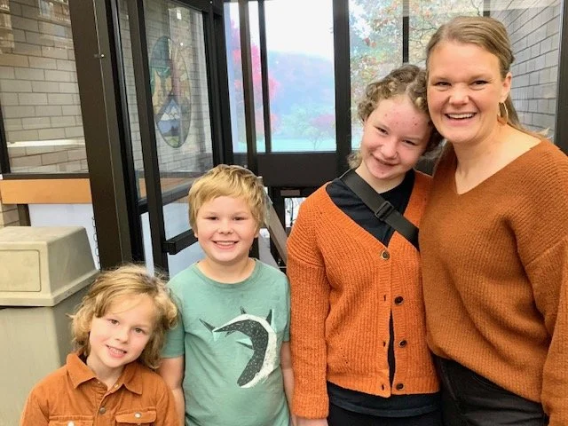 A woman with three children smiling indoors, with large windows and brick walls in the background.
