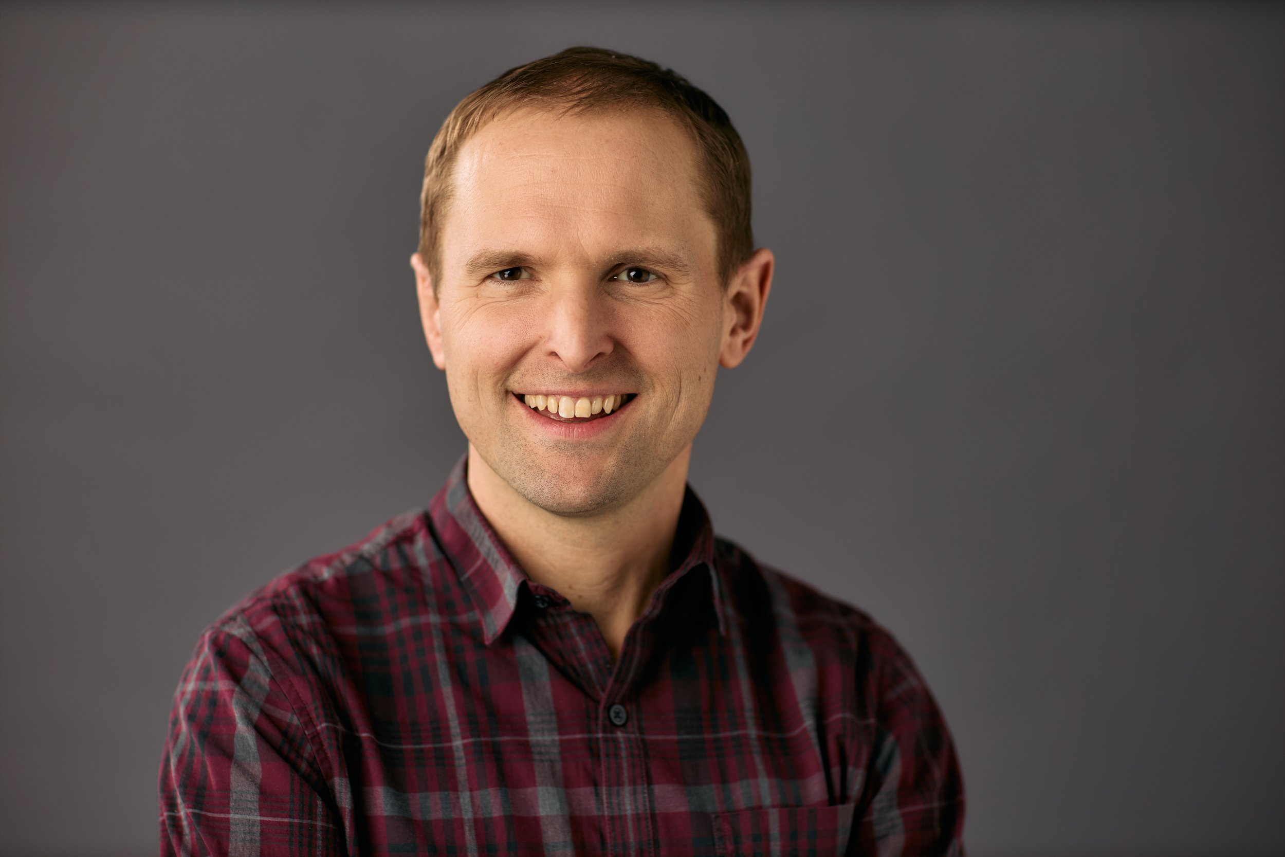 Portrait of a man smiling with short brown hair, wearing a red and black plaid shirt, against a plain dark gray background.