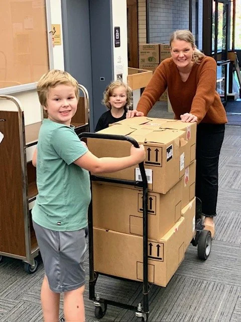 A woman and two children pose with a cart loaded with cardboard boxes indoors, smiling at the camera.