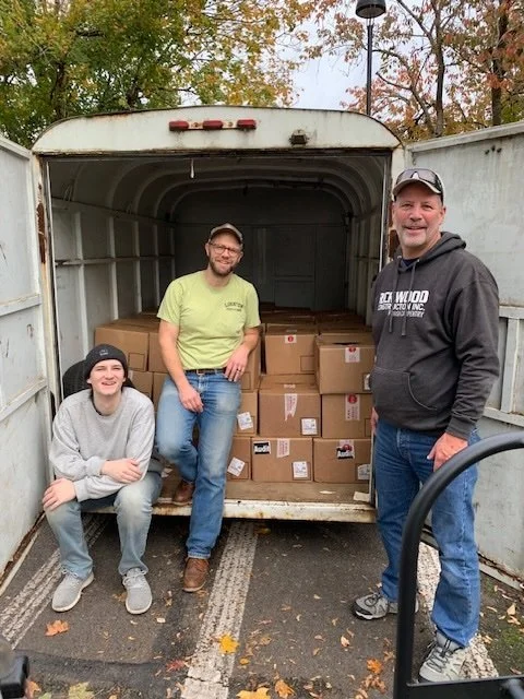 Three people posing in front of a truck filled with brown boxes, with autumn trees in the background.