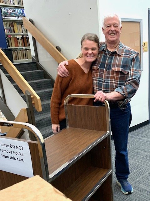 A woman and an elderly man smiling and hugging each other inside a library or bookstore. They are standing next to a rolling cart with a sign on it.