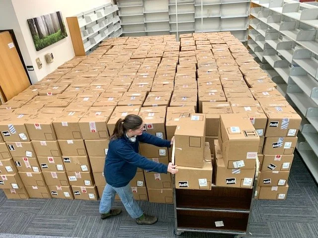 A woman organizing dozens of stacked cardboard boxes inside a retail store or warehouse.