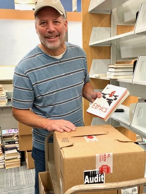 Smiling man wearing a beige cap and striped blue t-shirt in a bookstore, holding a book titled 'Ethics' while standing next to a cart with a cardboard box labeled 'Audit'.