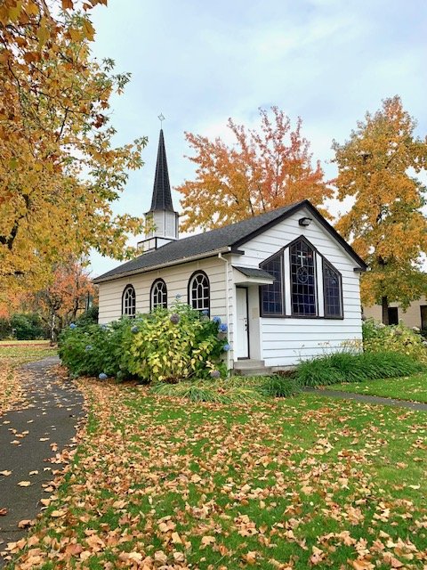 Small white church with black trim, large stained glass windows, and a steeple, surrounded by fall trees with orange and yellow leaves, some fallen on the sidewalk and grass.