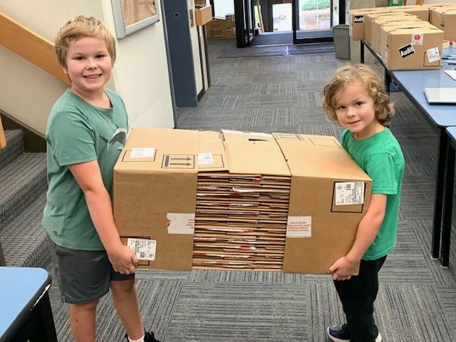 Two children, a boy and a girl, are holding a large cardboard box filled with envelopes inside a building with tables and boxes around.