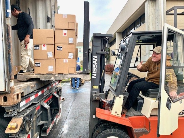 A man driving a forklift loading boxes onto a truck while another person stands on the truck. The boxes are labeled 'Audi'.