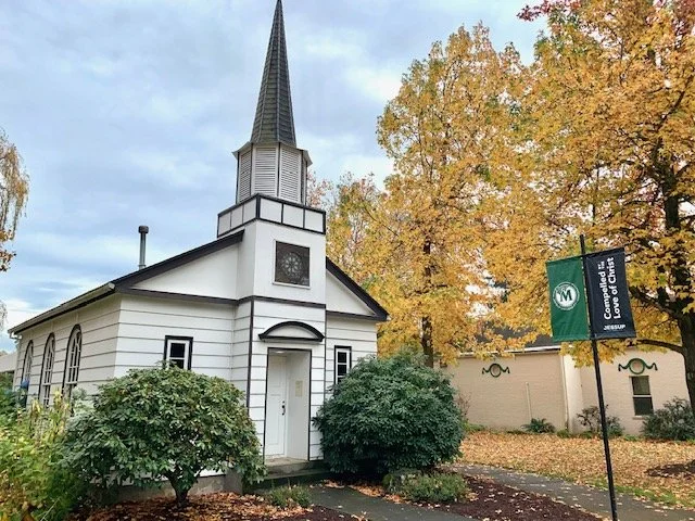 A small white church with a tall steeple, surrounded by green bushes and autumn trees with yellow leaves.