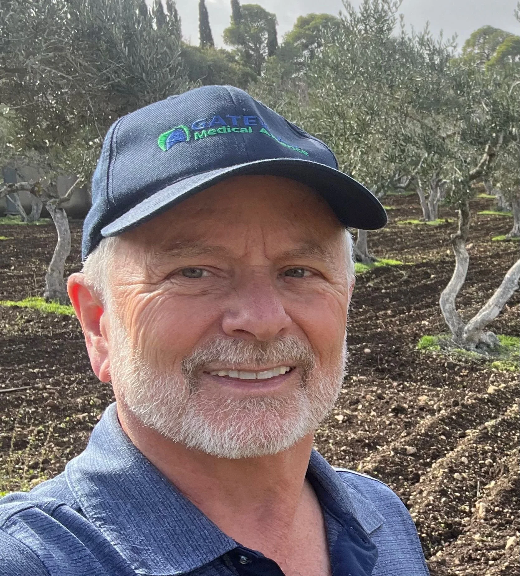 Close-up of a smiling man wearing a gray shirt and a navy blue cap with a medical association logo, standing in front of a landscape with trees and soil.