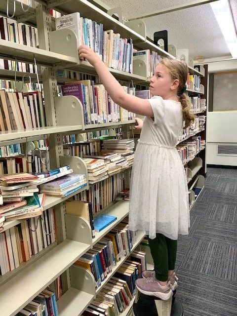 A young girl in a white dress and pink shoes is reaching for a book on a library shelf, standing on a small step stool.