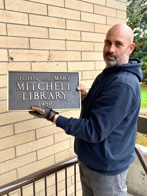 Man holding a plaque that reads 'John and Mary Mitchell Library 1980' outside a brick building.