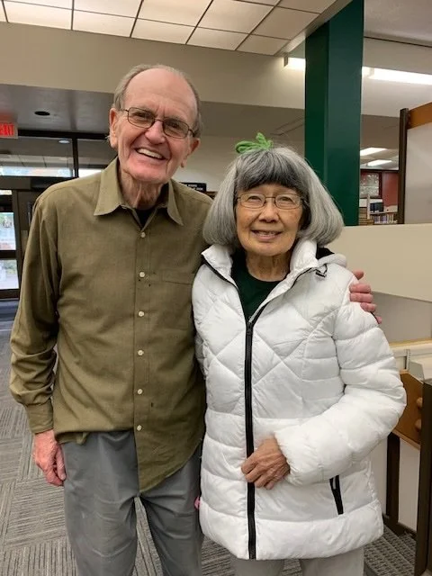 An elderly man and woman standing indoors, smiling and posing for the photo. The man is wearing glasses and a brown shirt, and the woman is wearing glasses and a white puffy jacket with a green bow in her hair.