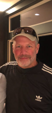 A man wearing a gray cap, sunglasses on his head, and a black Adidas jacket, smiling indoors near a window.