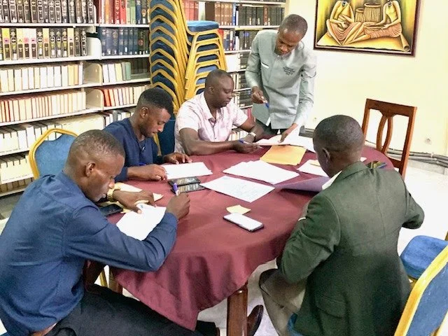Five men sit around a table with documents and electronic devices, in a room with bookshelves and artwork, while a woman stands and points at a sheet for discussion.