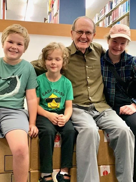 Four people, including three children and an adult man, sitting together in a library. They are smiling, with bookshelves visible in the background.