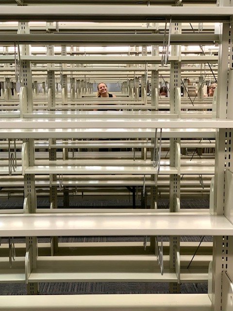 A person with a smiling face is seen behind empty store shelves in an aisle of a retail store.
