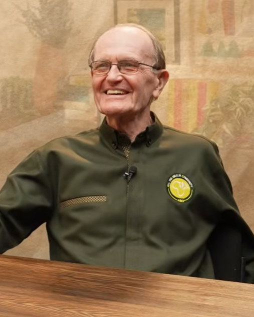 An elderly man with glasses, smiling, wearing a dark green jacket with a logo, seated at a wooden table.