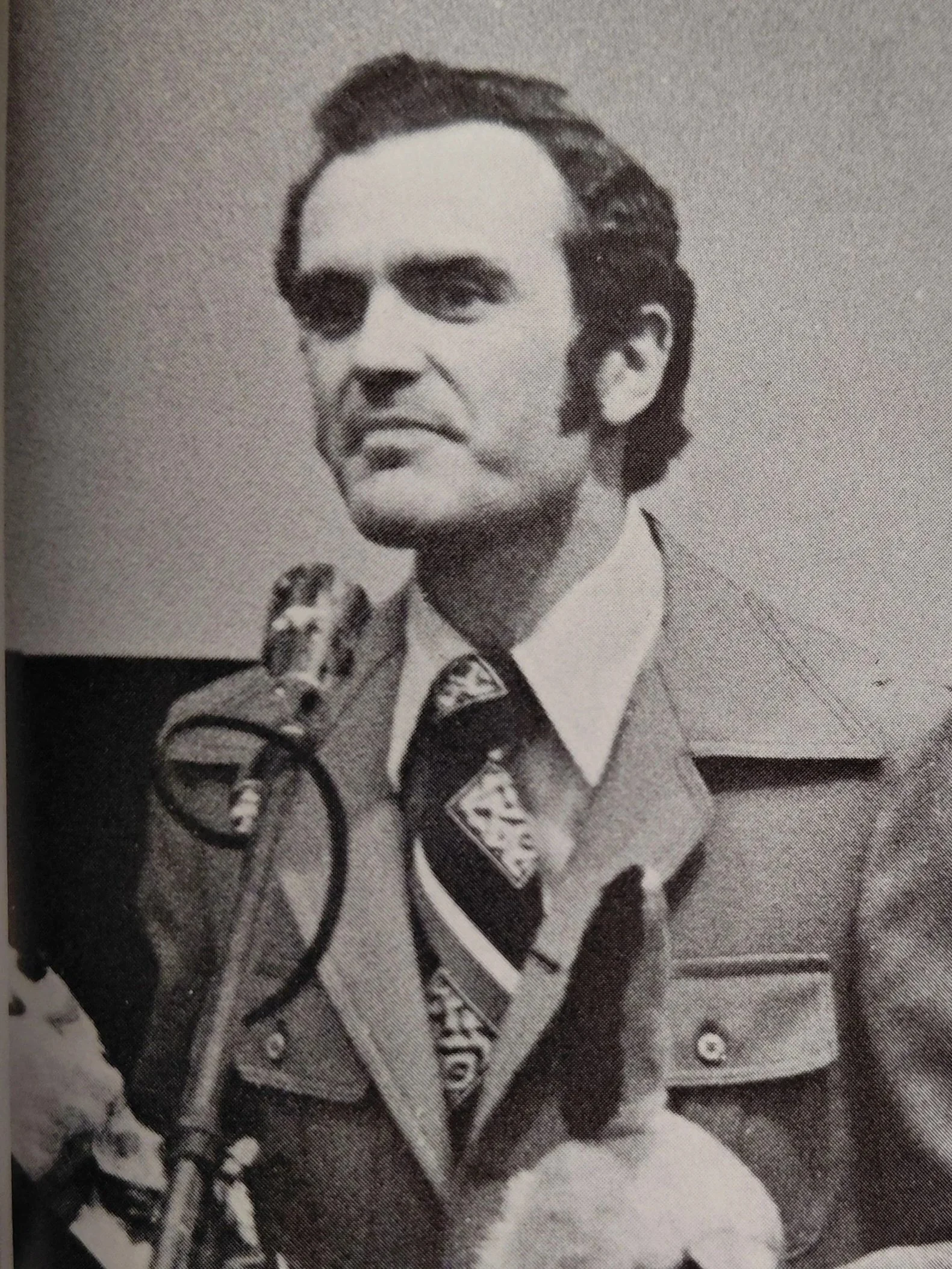 Black and white photo of a man in a military uniform with a patterned tie, sitting in front of a microphone, looking serious.