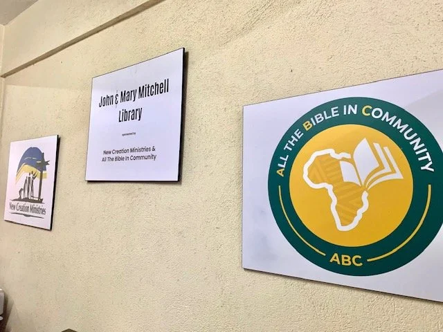 Signs on a beige wall for John & Mary Mitchell Library, New Creation Ministries, and All the Bible in Community (ABC).