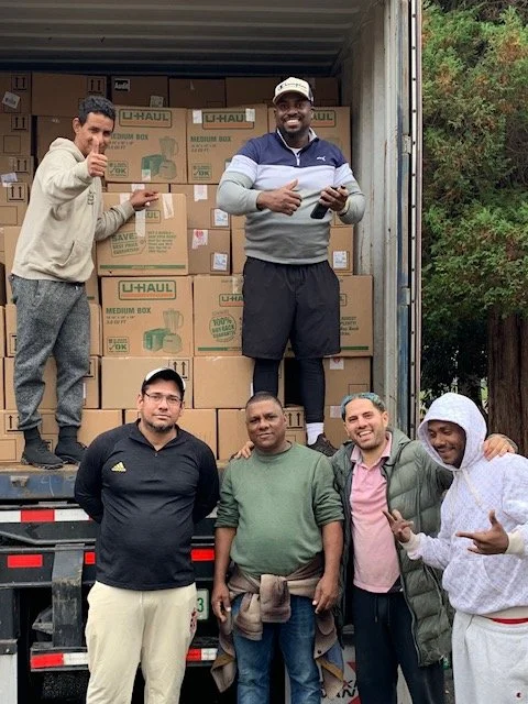 Six men standing in front of a truck filled with boxes, some on the ground and some stacked inside the truck. One man is standing on the edge of the truck's open door, smiling and giving a thumbs up. The others are standing in front, smiling, and mak