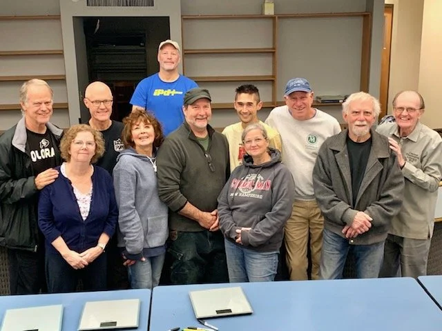 Group of ten people standing indoors, smiling, with a table in the foreground and empty shelves in the background.