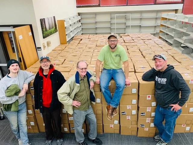 Five people standing in front of a wall of stacked cardboard boxes in a store or warehouse, smiling.