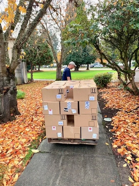 Man standing outdoors on a sidewalk next to a cart loaded with stacked cardboard boxes, with fallen autumn leaves surrounding the scene.