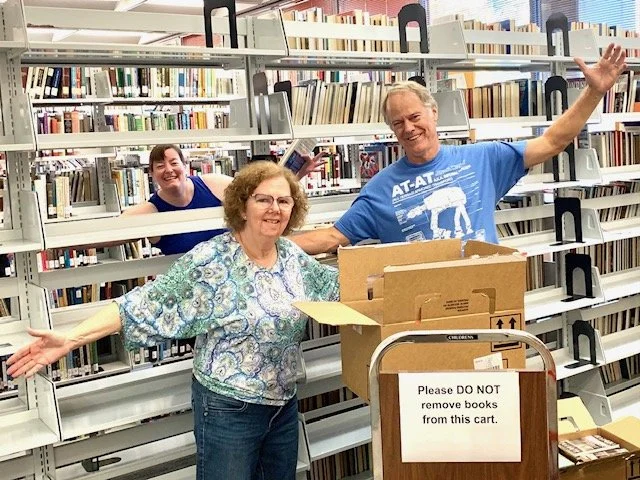 Three smiling people in a library with shelves filled with books. A woman and a man are next to a cart with a sign that says 'Please DO NOT remove books from this cart.' The woman has her arms outstretched, and the man is waving.