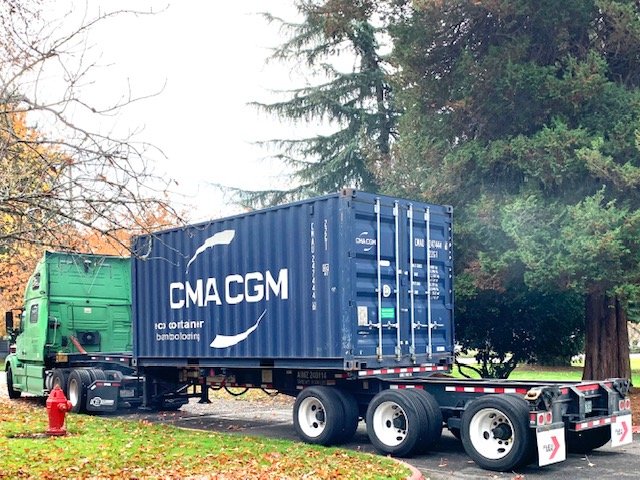 A large blue shipping container on a semi-truck trailer parked on a grassy area near trees.