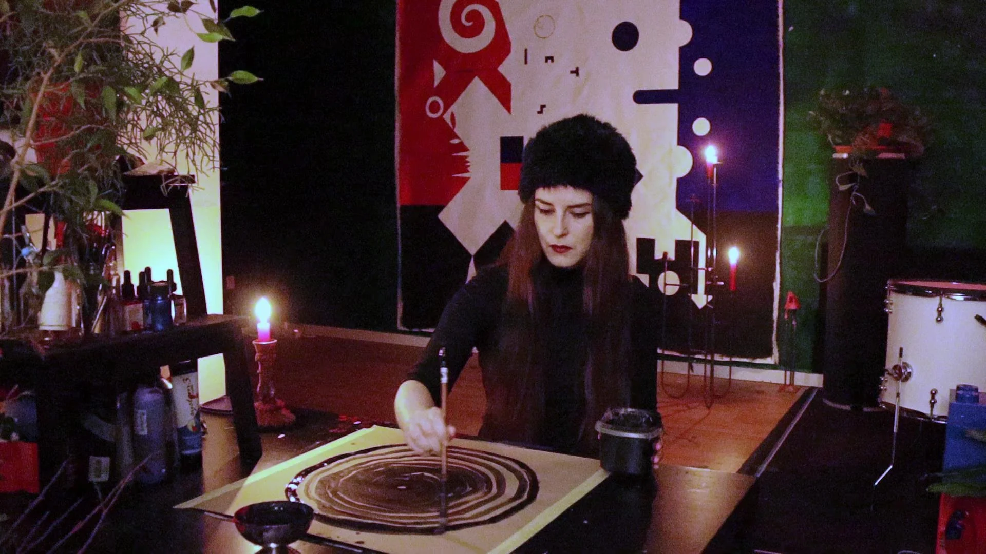 A woman with dark hair, wearing a black hat and black clothing, is painting a circular pattern on a large canvas with a brush. The room has a dark ambiance with candles, plants on the left, and abstract artwork on the wall in the background.