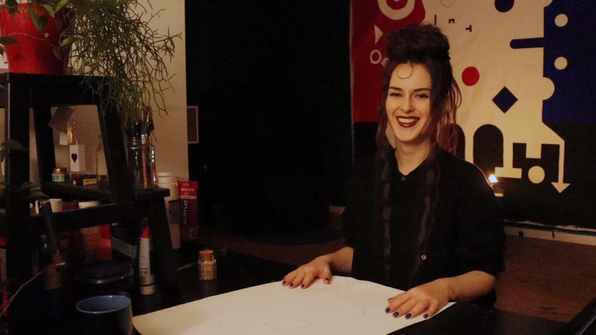 A woman with dark, curly hair styled in a half-updo, wearing a black shirt, is smiling and standing behind a table in a dimly lit room. There are art supplies on the table and a colorful, geometric-patterned wall in the background.