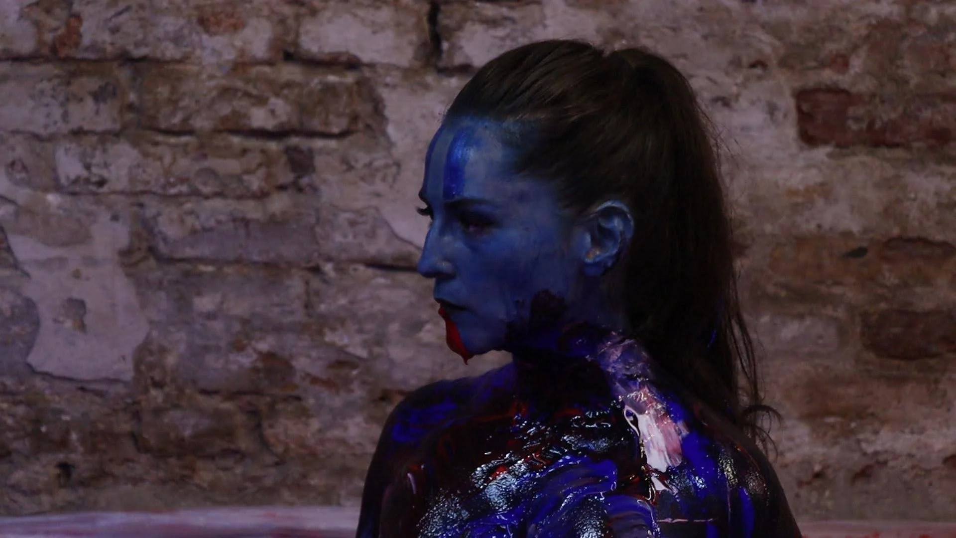 A woman with face and body covered in blue, red, and black paint, standing against a brick wall, in a side profile view.