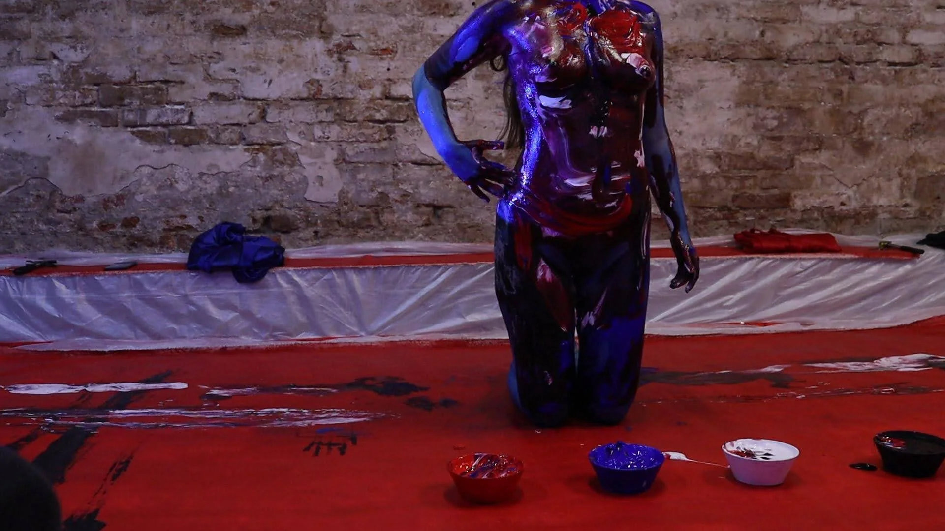 A person with their upper body and head painted in vibrant, swirling colors, kneeling on a red floor covered with paint, with a brick wall background. The person is touching their stomach with one hand, and their body is shiny with paint.