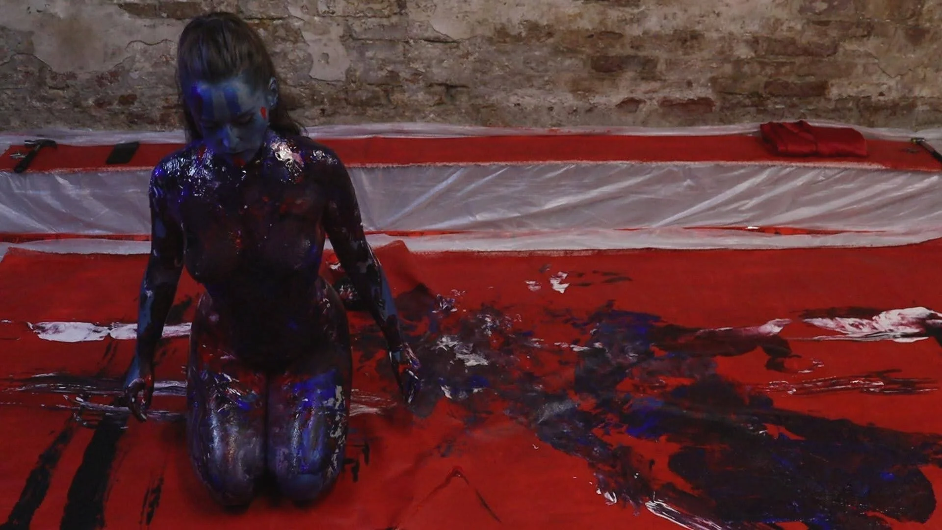 A person covered in black, blue, and white paint kneeling on a red canvas, with red and black paint splattered around them and an unfinished painting in the background.