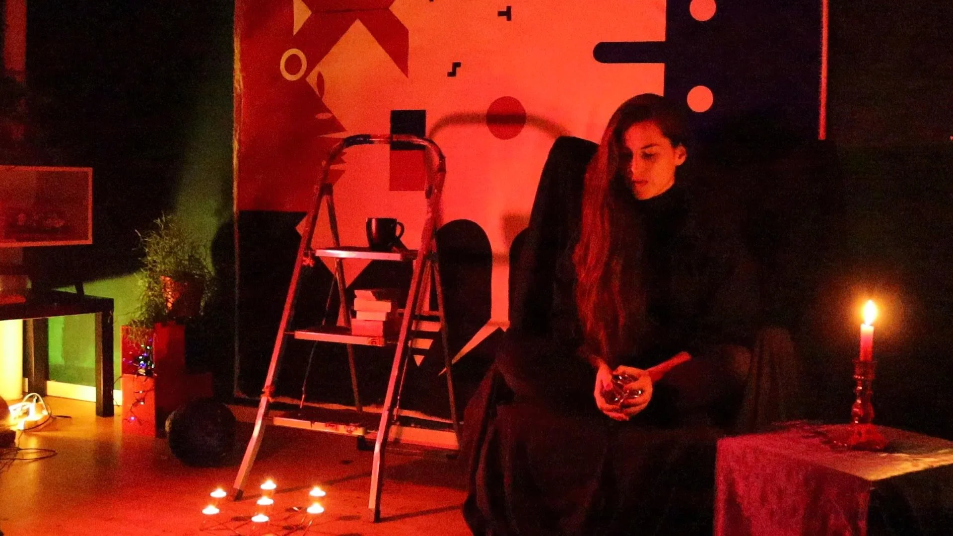 A woman with long hair sitting in a dimly lit room illuminated by a red candle, holding a glass, with a ladder shelf, books, a mug, and a potted plant in the background.