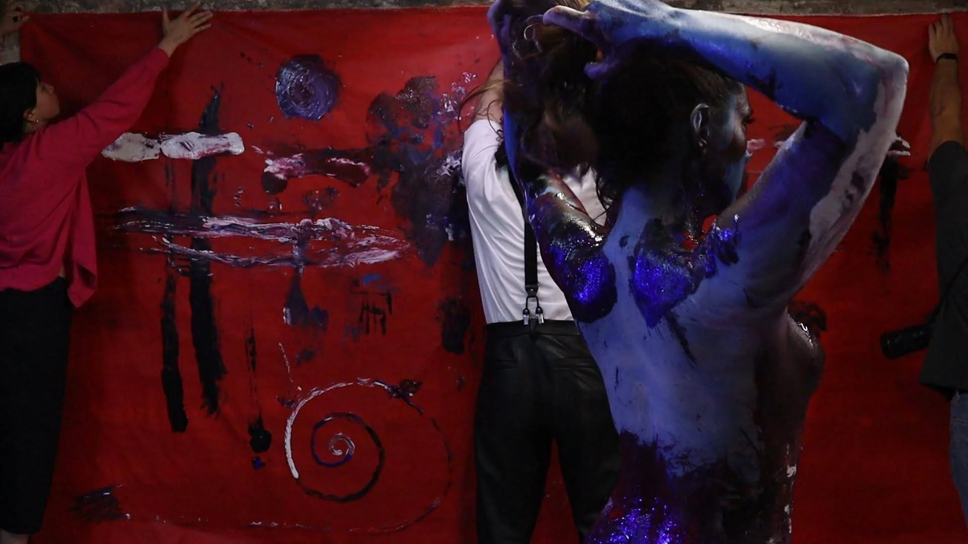 People creating abstract art on a large red canvas with paint and brushes.