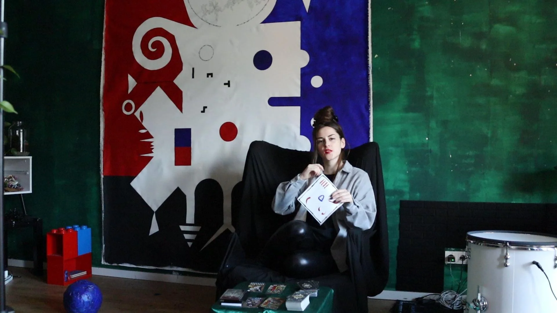 A woman with dark hair in a top knot seated in a black chair, holding a printed abstract face doodle, with a large abstract artwork behind her on a green wall. There are books, a blue globe, and a white drum in the room.