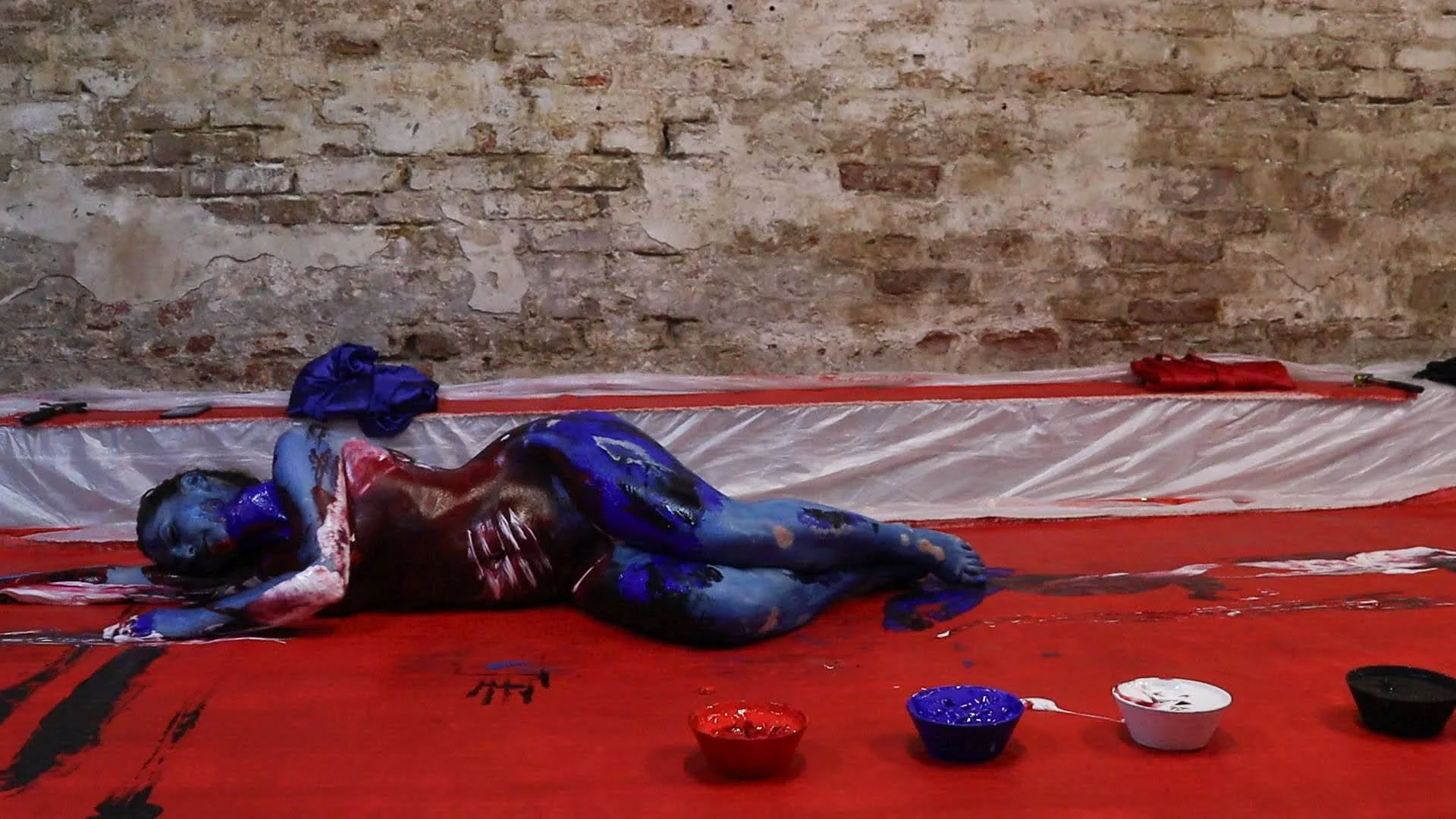 A person lying on the floor in front of a brick wall, covered in blue, red, and black paint, with art supplies and bowls of paint nearby.