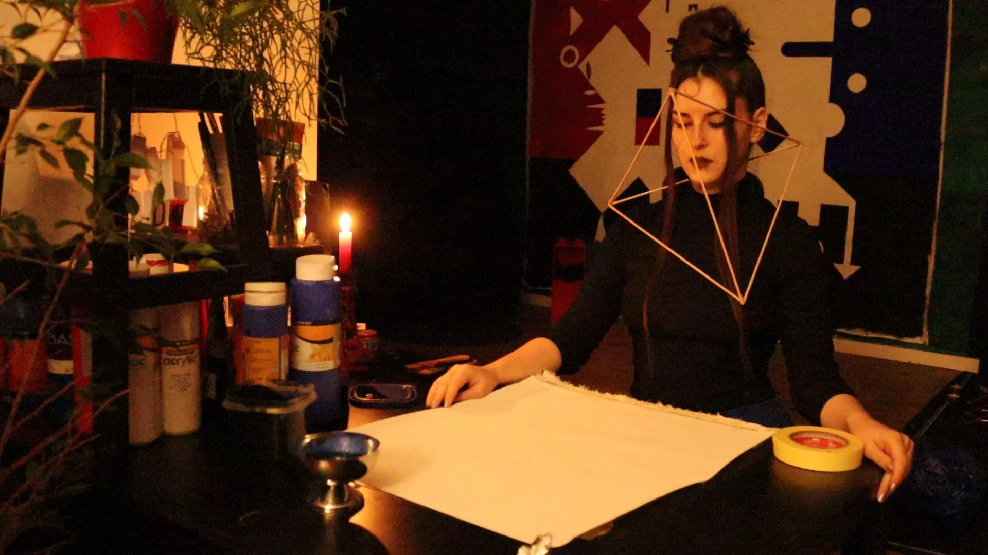 A woman with dark hair in a bun, wearing black clothing, sitting at a desk with art supplies, a candle, and tape. She is looking down at a large sheet of paper, with a geometric wire sculpture around her neck. The background features a colorful abstract poster.