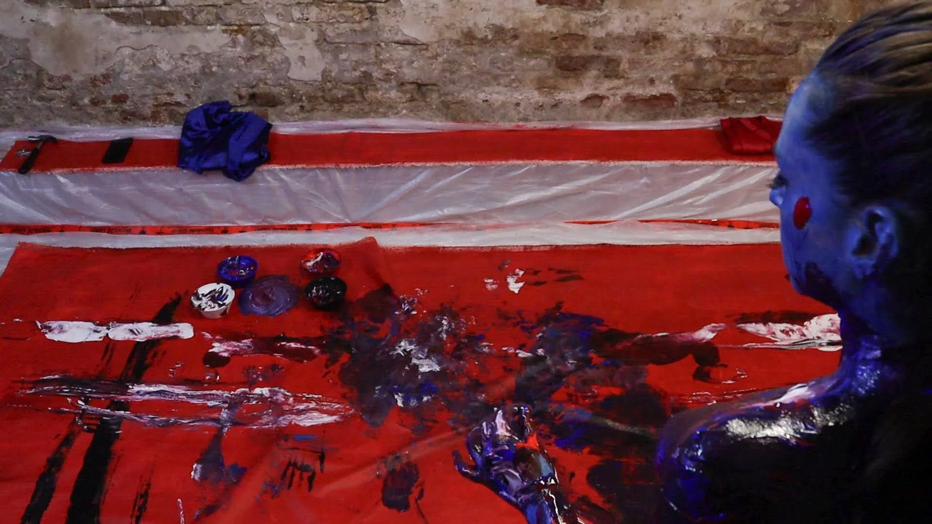 An artist's workspace with a red table covered in black, white, blue, and purple paint splatters and several small paint containers. A woman with her hair tied back is seen from the side, working on a painting in a room with brick walls and plastic-covered surfaces.