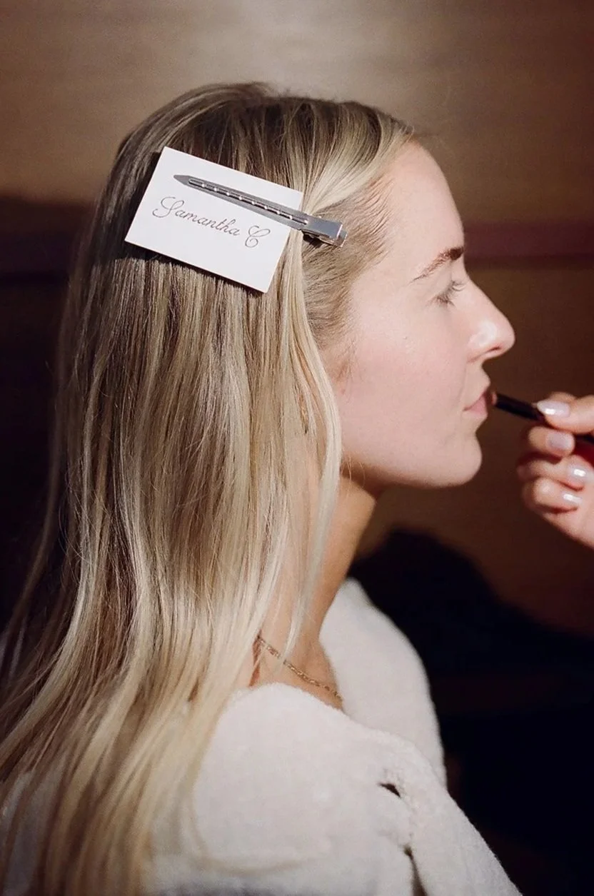 A woman with long blonde hair is getting her makeup done, with a name tag clipped to her hair saying "Samantha C". The woman has her eyes closed and someone is applying makeup to her lips.