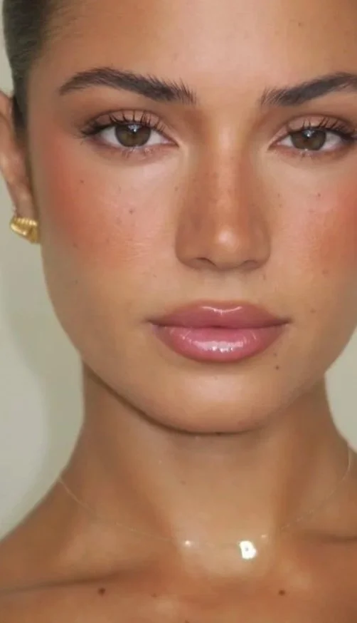 A close-up of a woman's face with light skin, brown eyes, well-groomed eyebrows, and glossy lips, wearing gold earrings and a delicate necklace.