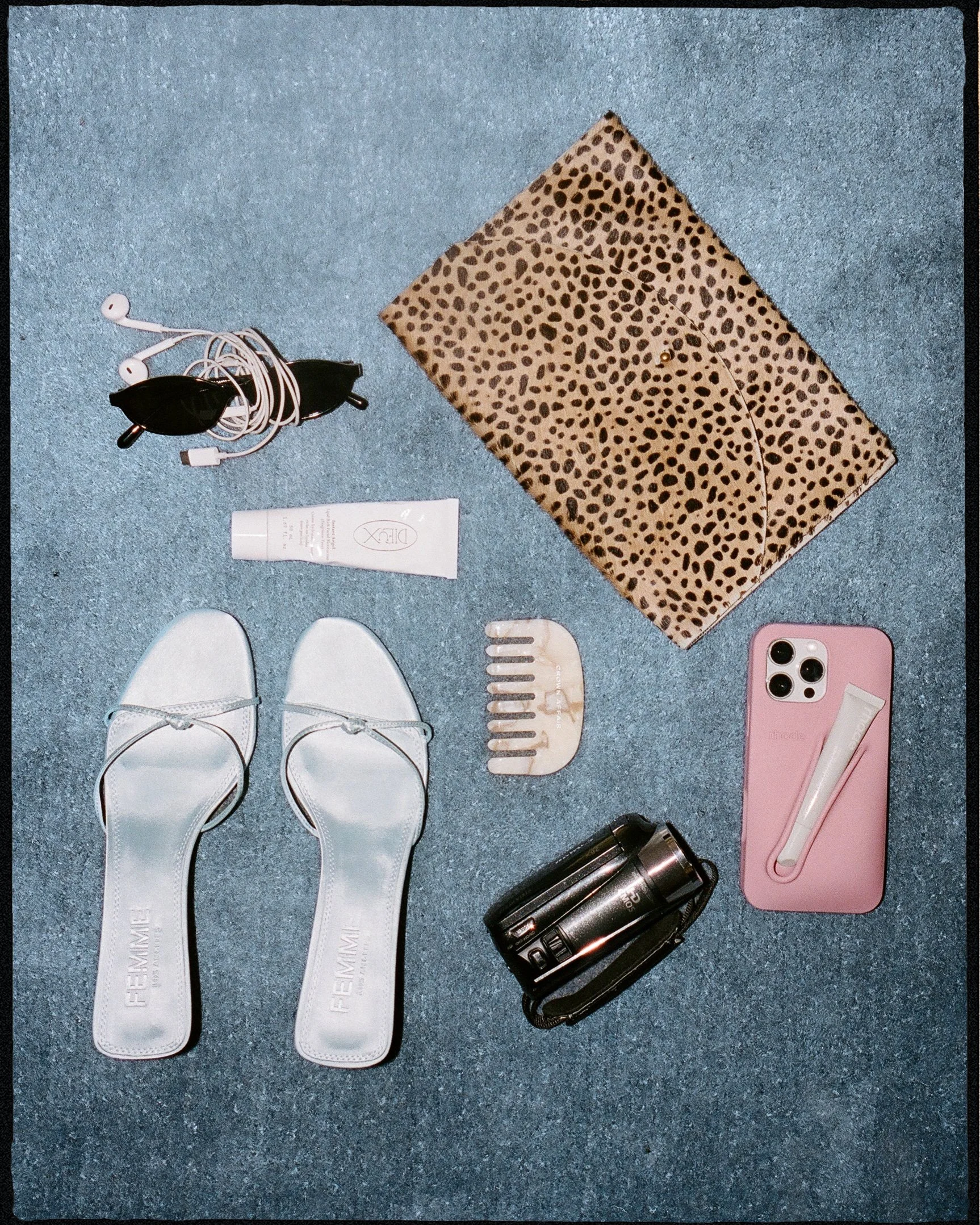 Flat lay of personal items on a blue carpet, including white sandals, a leopard print cloth, white earbuds, a tube of skincare cream, a pair of sunglasses, a pink phone with a white facial device, a black camera, and a beige hair comb.