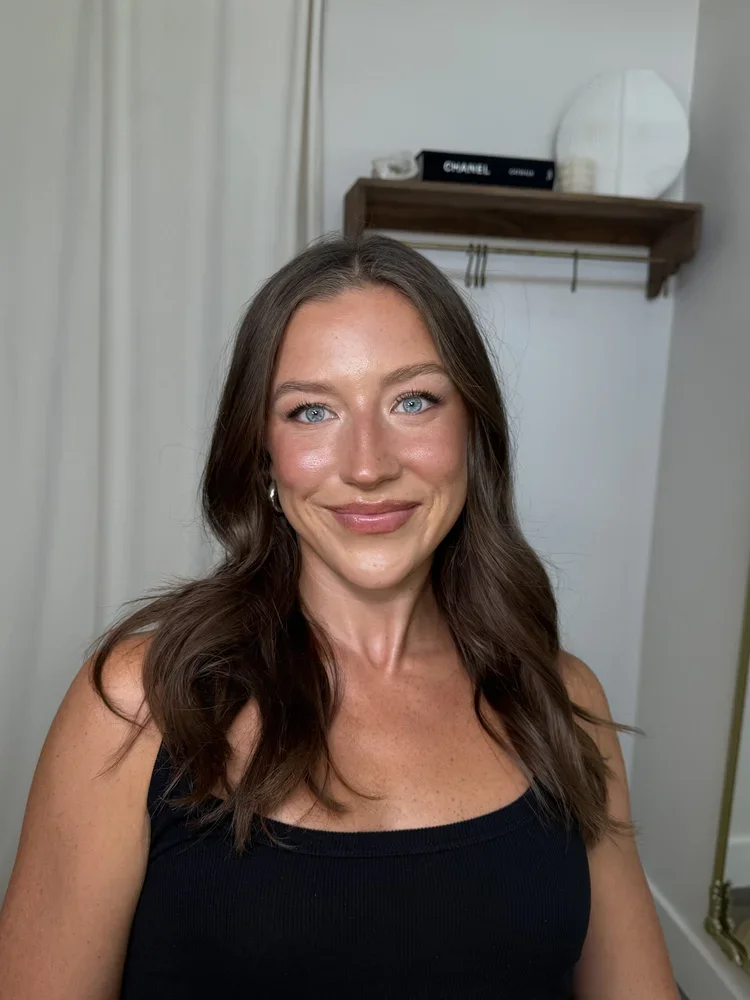 A woman with long wavy brown hair and blue eyes smiling in a black tank top, with a background showing a wooden shelf and white curtains.