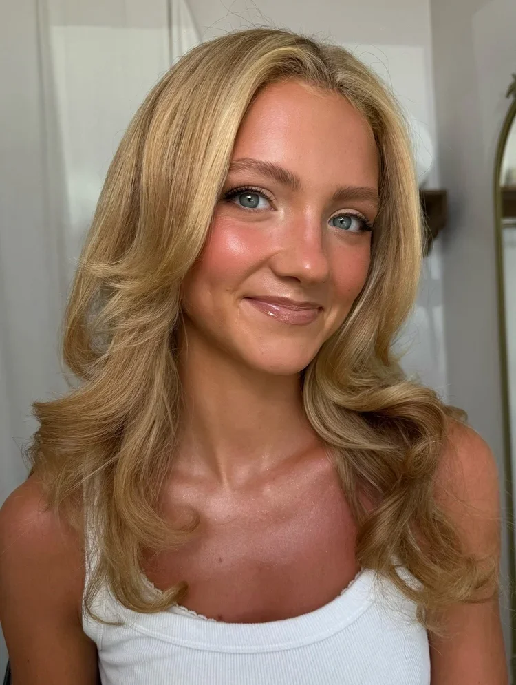 A young woman with long, wavy blonde hair, blue eyes, and light skin, smiling in a white tank top, standing indoors in front of a mirror.