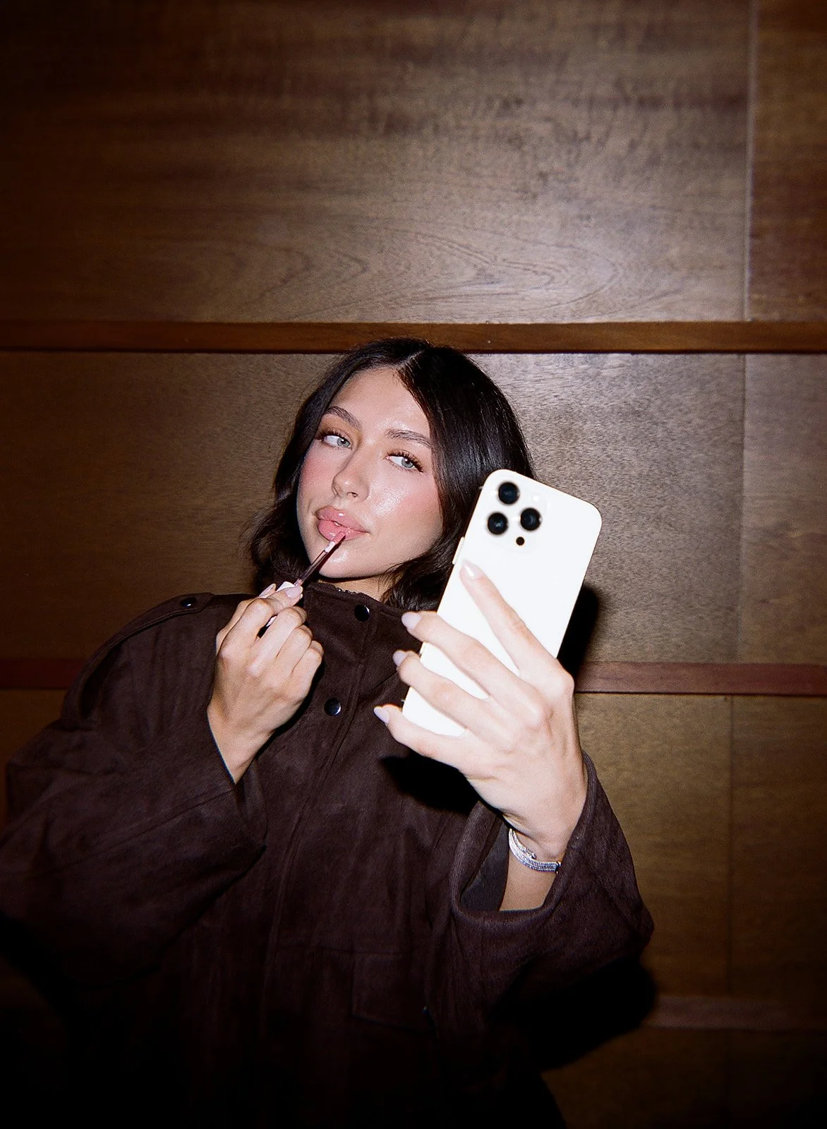 A woman taking a selfie with a smartphone while applying lip gloss.